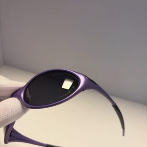 K&ouml;nnte beinhalten: Violette Sonnenbrille mit dunklen Gl&auml;sern. Die Sonnenbrille hat ein schlankes, modernes Design mit einem geschwungenen Rahmen und schwarzen Akzenten an den B&uuml;geln. Auf den Gl&auml;sern ist das Wort "PRIZM" aufgedruckt.