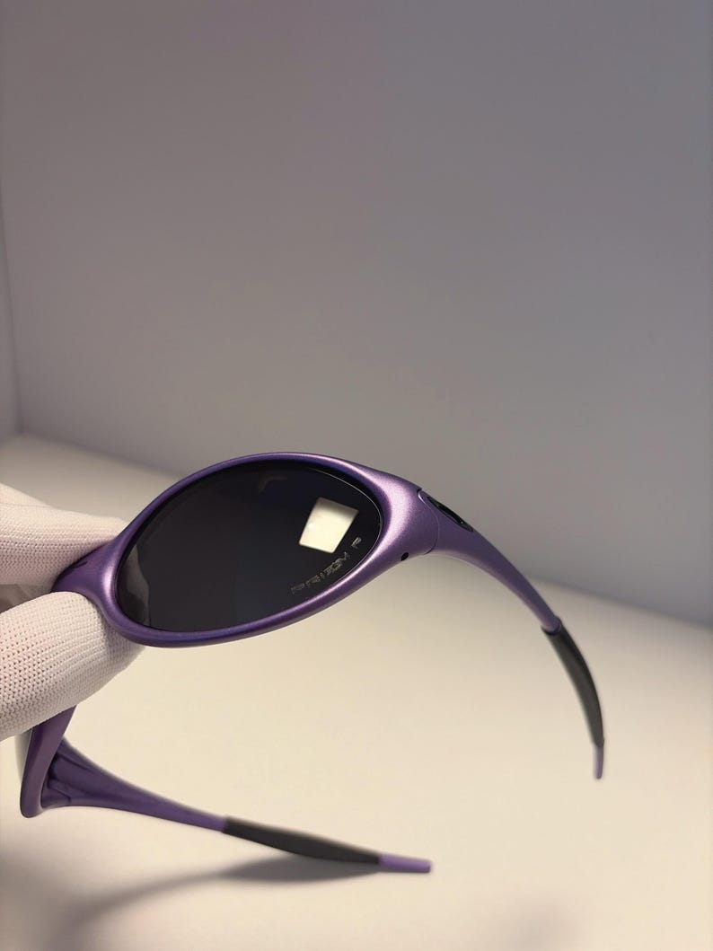 K&ouml;nnte beinhalten: Violette Sonnenbrille mit dunklen Gl&auml;sern. Die Sonnenbrille hat ein schlankes, modernes Design mit einem geschwungenen Rahmen und schwarzen Akzenten an den B&uuml;geln. Auf den Gl&auml;sern ist das Wort "PRIZM" aufgedruckt.