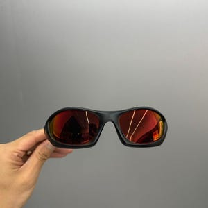 May include: Black sunglasses with red mirrored lenses. The sunglasses have a rounded, sporty design. The lenses reflect the surrounding environment. The frame is a matte black colour.