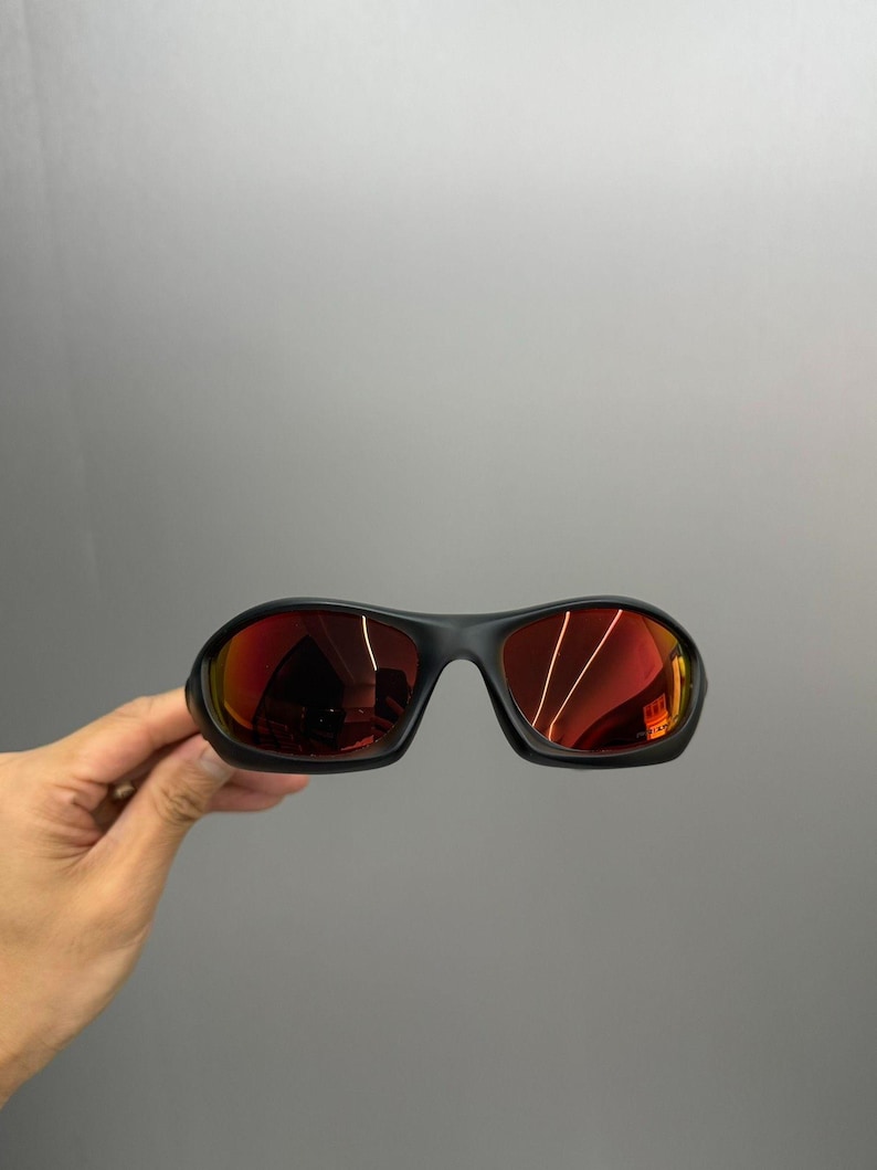 May include: Black sunglasses with red mirrored lenses. The sunglasses have a rounded, sporty design. The lenses reflect the surrounding environment. The frame is a matte black colour.