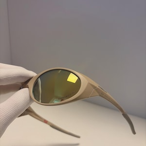 May include: Tan-colored sunglasses with a curved design and mirrored lenses. The sunglasses have a sleek, modern look with a subtle logo on the lens. The arms of the sunglasses are a lighter shade of tan and have a curved design.
