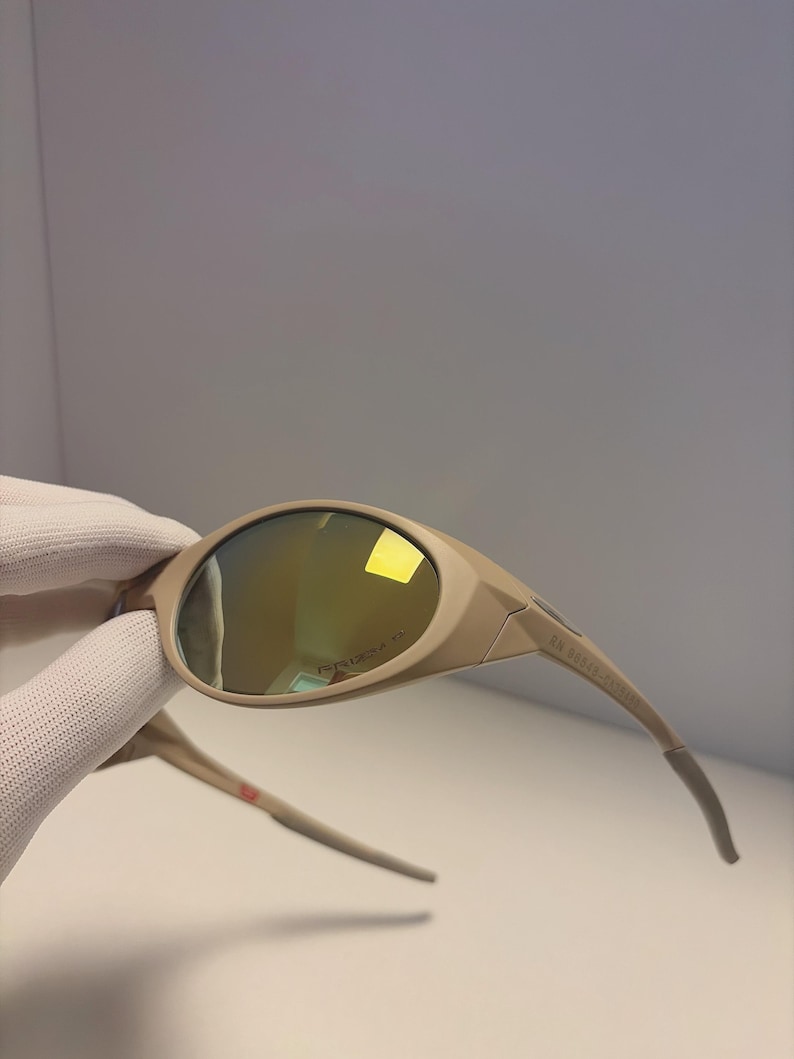 May include: Tan-colored sunglasses with a curved design and mirrored lenses. The sunglasses have a sleek, modern look with a subtle logo on the lens. The arms of the sunglasses are a lighter shade of tan and have a curved design.