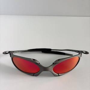 May include: Gray sunglasses with red lenses. The sunglasses have a sleek, modern design with a thin frame and curved temples. The lenses are a vibrant red color, and the frame is a matte gray.