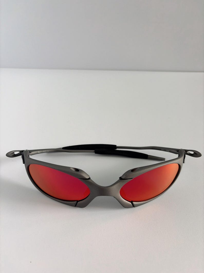 May include: Gray sunglasses with red lenses. The sunglasses have a sleek, modern design with a thin frame and curved temples. The lenses are a vibrant red color, and the frame is a matte gray.