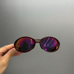May include: Brown-framed sunglasses with oval lenses. The lenses reflect a vibrant gradient of pink and purple hues. The sunglasses have a classic, rounded shape, suitable for various face shapes. The frame appears to be made of a durable material.