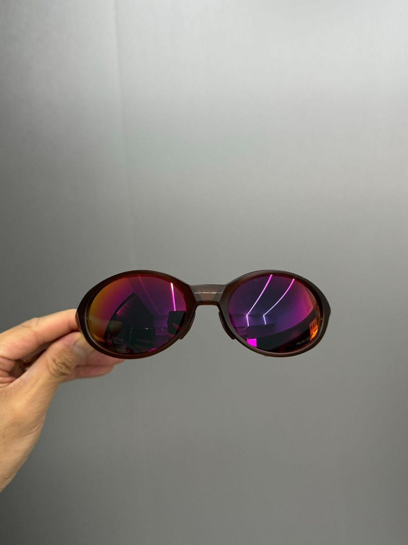 Puede incluir: Gafas de sol con montura marr&oacute;n y lentes ovaladas. Las lentes reflejan un vibrante degradado de tonos rosas y morados. Las gafas de sol tienen una forma cl&aacute;sica y redondeada, adecuada para varias formas de rostro. La montura parece estar hecha de un material duradero.