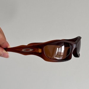 May include: Brown sunglasses with a curved frame and tinted lenses. The sunglasses have a small silver emblem on the side. The lenses reflect the surrounding environment.