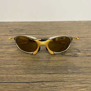 May include: A pair of sporty sunglasses with a silver frame and yellow accents. The lenses are a warm brown color, and the design is sleek and modern. The sunglasses are resting on a wooden surface.