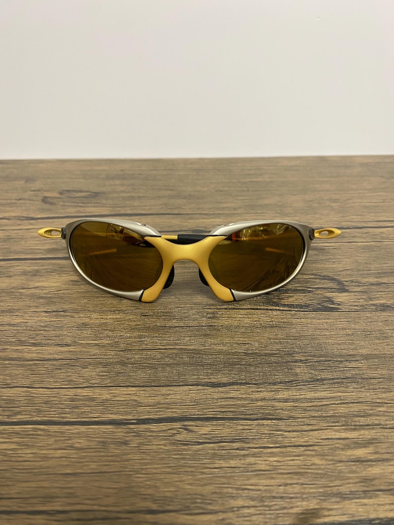 May include: A pair of sporty sunglasses with a silver frame and yellow accents. The lenses are a warm brown color, and the design is sleek and modern. The sunglasses are resting on a wooden surface.