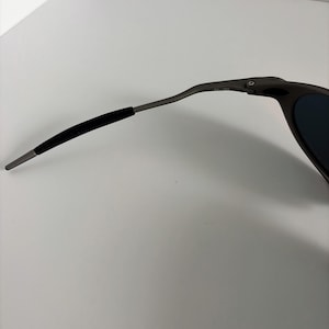 May include: Close-up of a pair of dark gray sunglasses. The sunglasses have a sleek, modern design with a curved frame and dark lenses. The arm of the glasses has a black rubber grip for comfort.