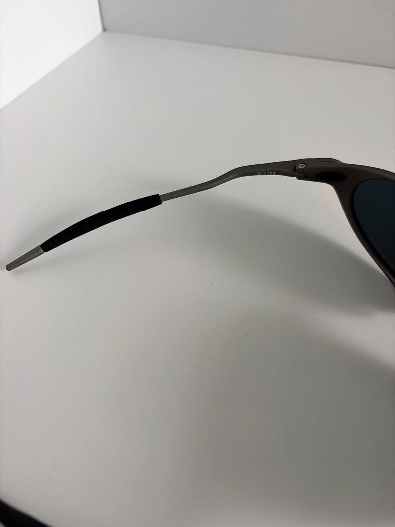 May include: Close-up of a pair of dark gray sunglasses. The sunglasses have a sleek, modern design with a curved frame and dark lenses. The arm of the glasses has a black rubber grip for comfort.