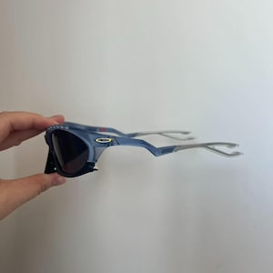 May include: Blue sunglasses with dark lenses and silver arms. The sunglasses have a distinctive, angular frame design. The lenses are dark, and the frame is a light blue colour. The arms are silver and curve around the ears.