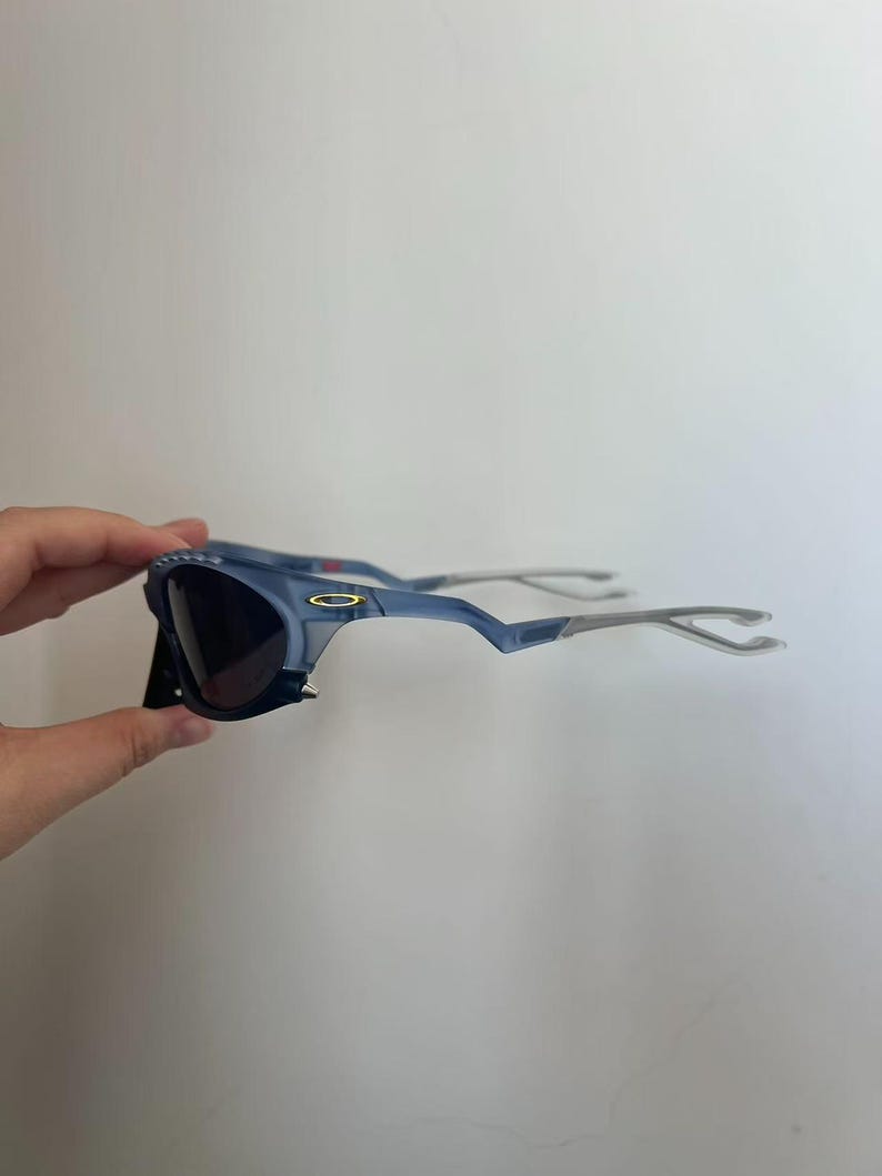 May include: Blue sunglasses with dark lenses and silver arms. The sunglasses have a distinctive, angular frame design. The lenses are dark, and the frame is a light blue colour. The arms are silver and curve around the ears.