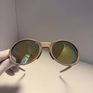 May include: Tan-colored sunglasses with mirrored lenses. The sunglasses have a rounded frame and a sleek, modern design. The lenses reflect a golden hue. The arms of the sunglasses are thin and extend outward. The sunglasses are held by a gloved hand.