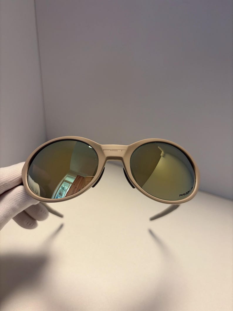 May include: Tan-colored sunglasses with mirrored lenses. The sunglasses have a rounded frame and a sleek, modern design. The lenses reflect a golden hue. The arms of the sunglasses are thin and extend outward. The sunglasses are held by a gloved hand.