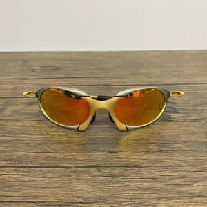 Puede incluir: Gafas de sol con montura dorada y lentes espejadas naranjas. Las gafas de sol tienen un dise&ntilde;o elegante y moderno, con una forma curvada y un puente fino. Las lentes reflejan la luz ambiental.