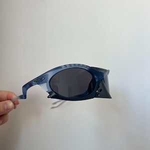 May include: Blue and grey sunglasses with a distinctive, angular frame design. The lenses are dark, providing shade. The sunglasses are held by a hand against a white background.