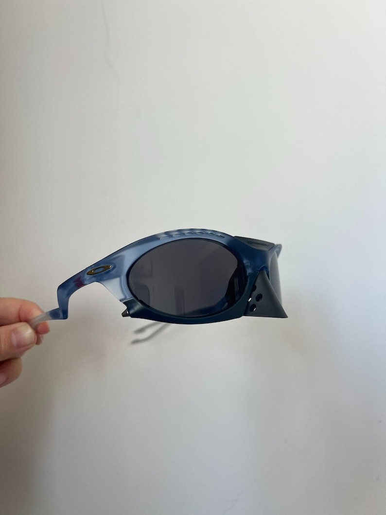 May include: Blue and grey sunglasses with a distinctive, angular frame design. The lenses are dark, providing shade. The sunglasses are held by a hand against a white background.