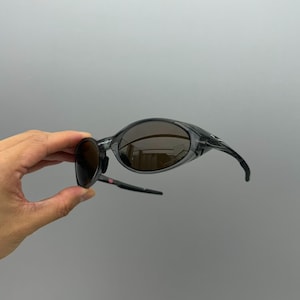 May include: A pair of wraparound sunglasses with a transparent gray frame and dark brown lenses. The sunglasses have a sporty design with curved temples and a sleek, modern aesthetic.
