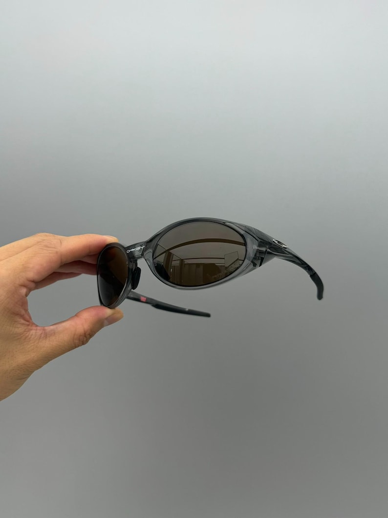 May include: A pair of wraparound sunglasses with a transparent gray frame and dark brown lenses. The sunglasses have a sporty design with curved temples and a sleek, modern aesthetic.