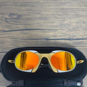 Puede incluir: Gafas de sol doradas y plateadas con lentes de color naranja brillante dentro de un estuche negro abierto. Las gafas tienen un dise&ntilde;o de montura curvada &uacute;nico. El estuche se encuentra sobre un fondo de madera.