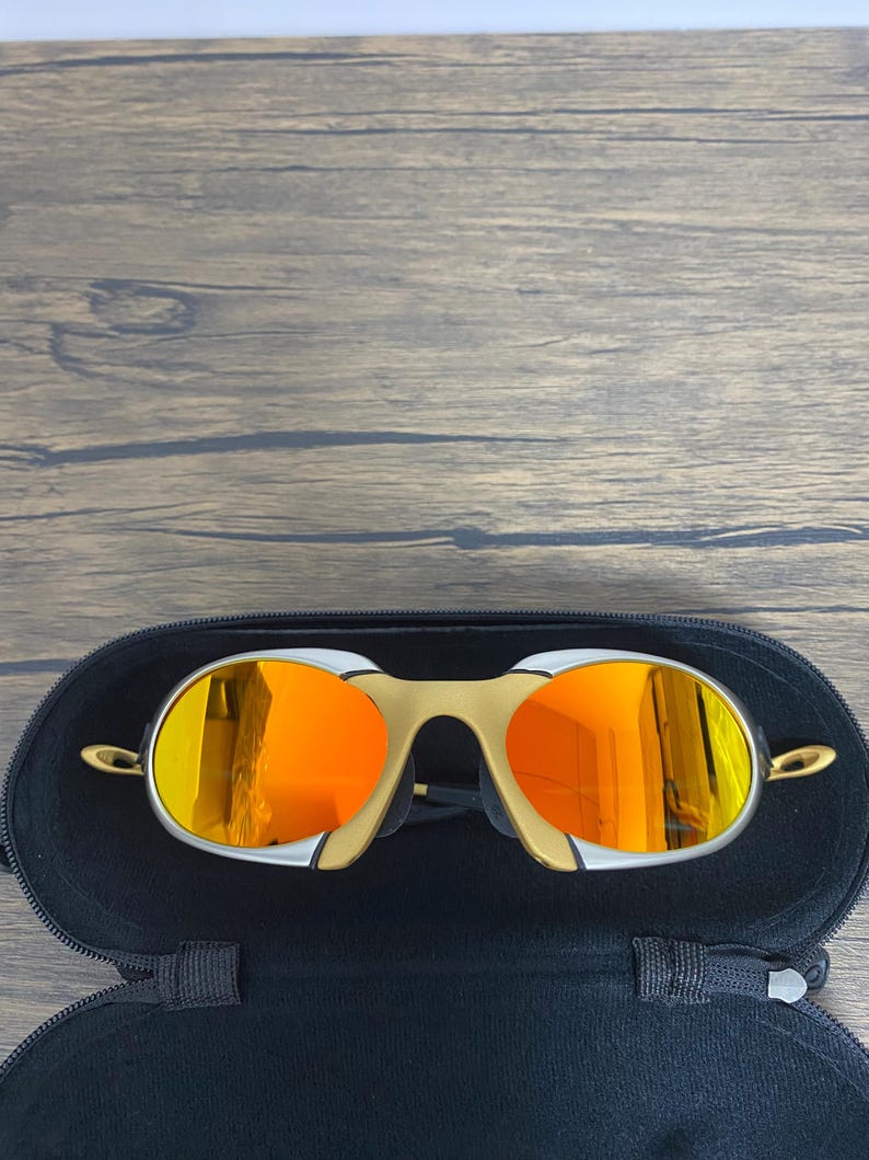Puede incluir: Gafas de sol doradas y plateadas con lentes de color naranja brillante dentro de un estuche negro abierto. Las gafas tienen un dise&ntilde;o de montura curvada &uacute;nico. El estuche se encuentra sobre un fondo de madera.