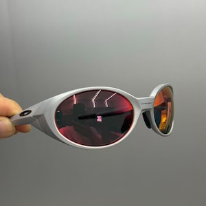 Puede incluir: Gafas de sol plateadas con lentes tintadas en rojo. Las lentes ovaladas est&aacute;n engastadas en una montura moderna. Las gafas de sol tienen un dise&ntilde;o deportivo, adecuado para actividades al aire libre.