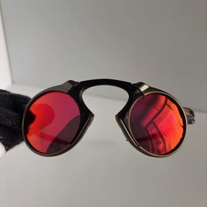 May include: Sunglasses with a black bridge and dark metal frames. The lenses are a vibrant red color. The sunglasses have a unique, modern design.