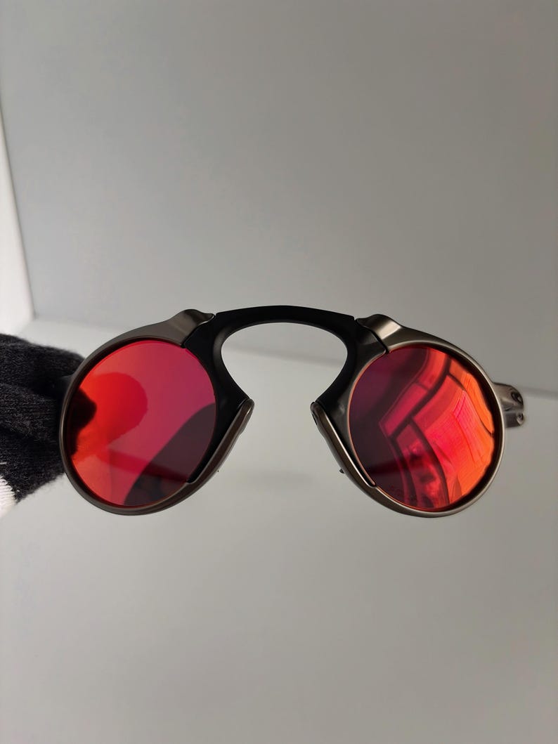 May include: Sunglasses with a black bridge and dark metal frames. The lenses are a vibrant red color. The sunglasses have a unique, modern design.