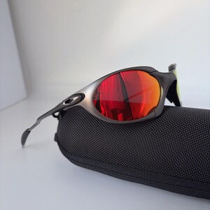 May include: Sport sunglasses with a black frame and red-orange lenses rest on a black textured case. The sunglasses have a sleek, modern design, with a silver accent on the frame.