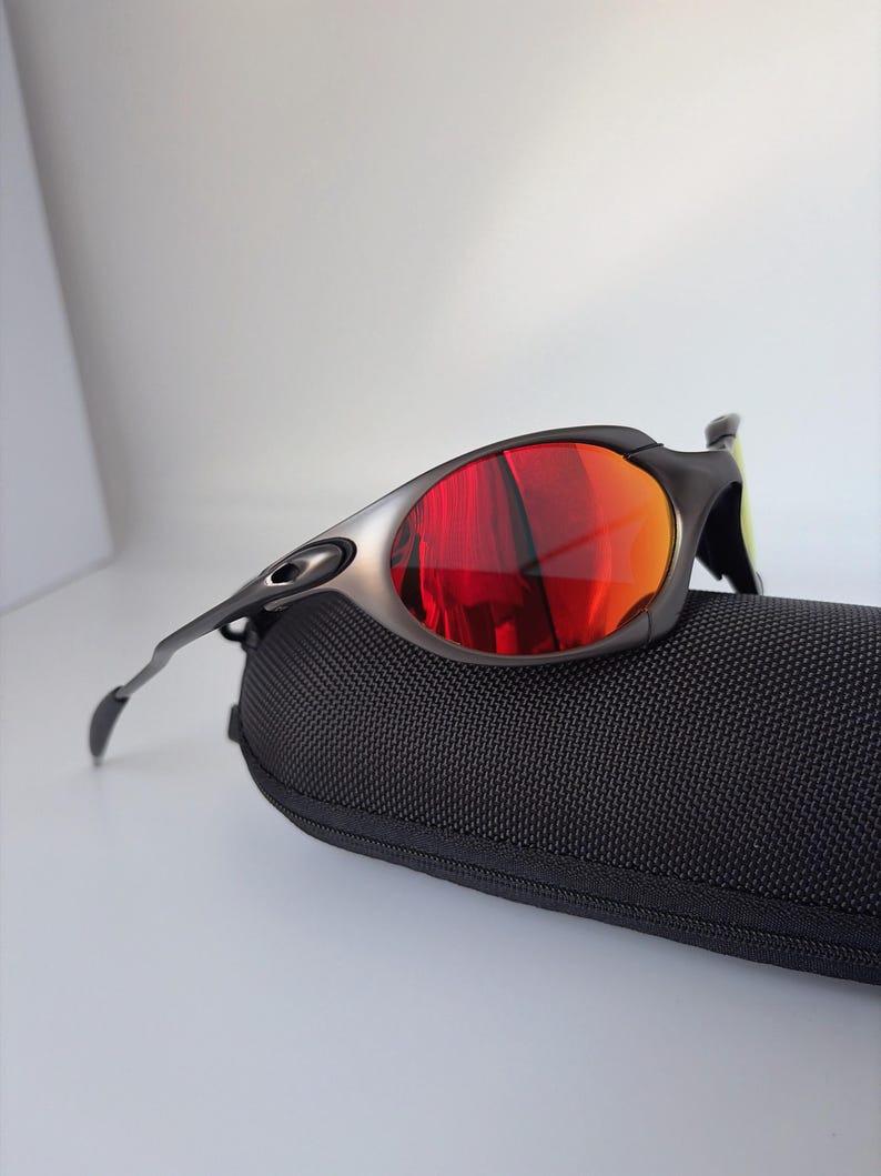 May include: Sport sunglasses with a black frame and red-orange lenses rest on a black textured case. The sunglasses have a sleek, modern design, with a silver accent on the frame.
