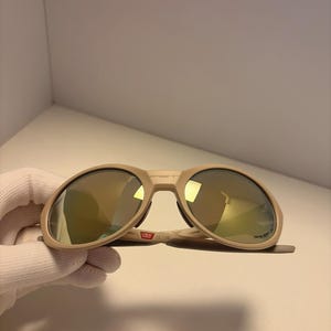 May include: Tan sunglasses with gold mirrored lenses. The sunglasses have a thick frame and a bridge with the text "USA" printed on it. The lenses are oval-shaped.
