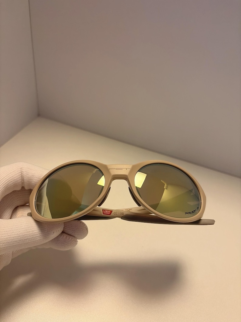 May include: Tan sunglasses with gold mirrored lenses. The sunglasses have a thick frame and a bridge with the text "USA" printed on it. The lenses are oval-shaped.