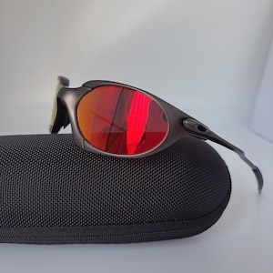 May include: Dark grey sunglasses with red mirrored lenses sit on a black textured case. The sunglasses have a sleek, modern design with a curved frame and a small, yellow-tinted lens on the side.