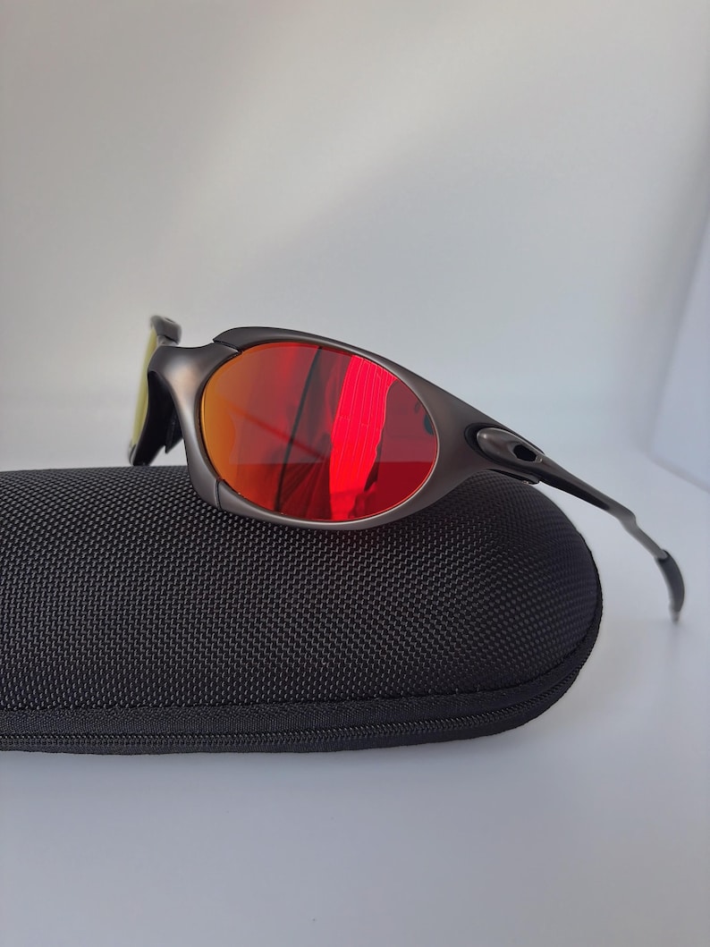 May include: Dark grey sunglasses with red mirrored lenses sit on a black textured case. The sunglasses have a sleek, modern design with a curved frame and a small, yellow-tinted lens on the side.
