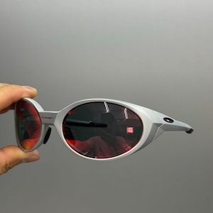 Puede incluir: Gafas de sol plateadas con lentes ovaladas rojas. El dise&ntilde;o es moderno y curvado, con un peque&ntilde;o logotipo en el lateral. Las lentes rojas son vibrantes y llamativas.