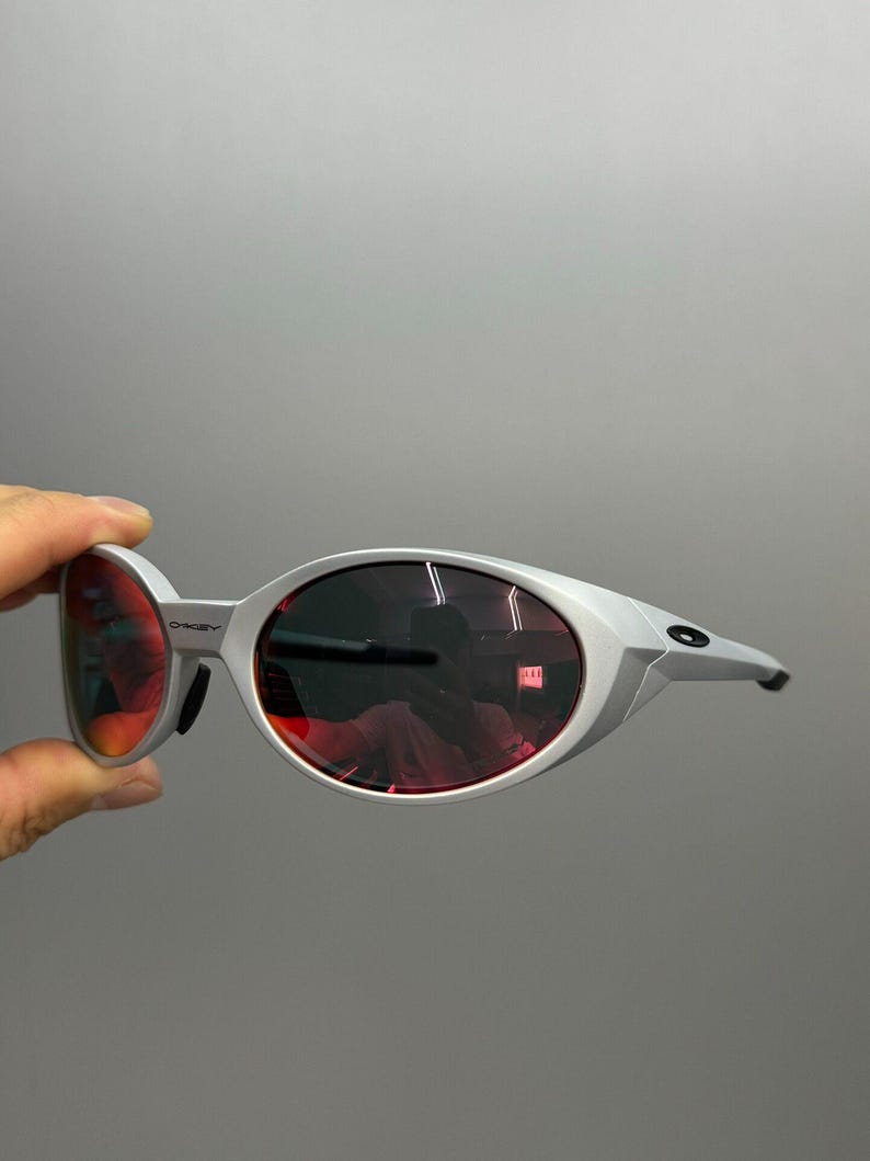 Puede incluir: Gafas de sol plateadas con lentes ovaladas rojas. El dise&ntilde;o es moderno y curvado, con un peque&ntilde;o logotipo en el lateral. Las lentes rojas son vibrantes y llamativas.