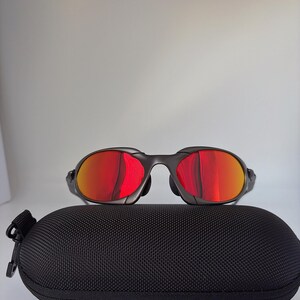 May include: Black sunglasses with red lenses sit on a black, zipped case. The sunglasses have a sleek, modern design with a dark frame. The lenses reflect a bright red hue, contrasting with the black case.