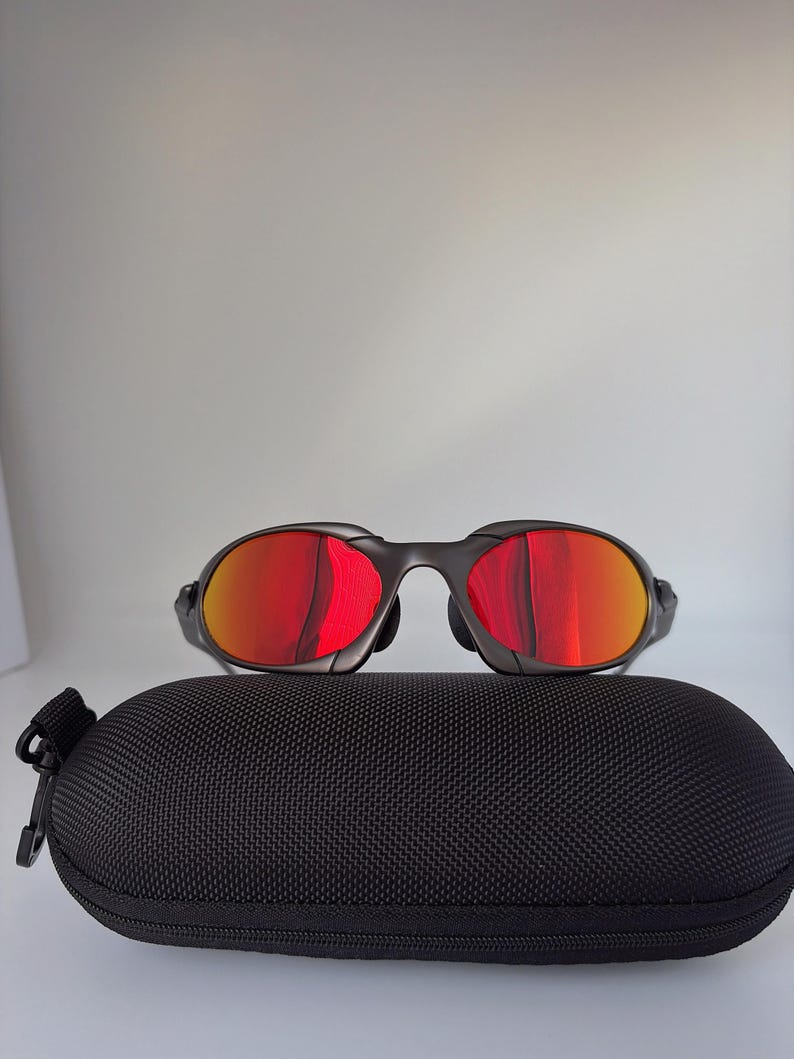 May include: Black sunglasses with red lenses sit on a black, zipped case. The sunglasses have a sleek, modern design with a dark frame. The lenses reflect a bright red hue, contrasting with the black case.