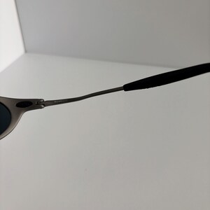 May include: Close-up of a pair of sunglasses with a matte gray frame and dark lenses. The temples are thin and curve towards the ear, with black rubberized tips for comfort. The text "ROMEO" is visible on the frame.