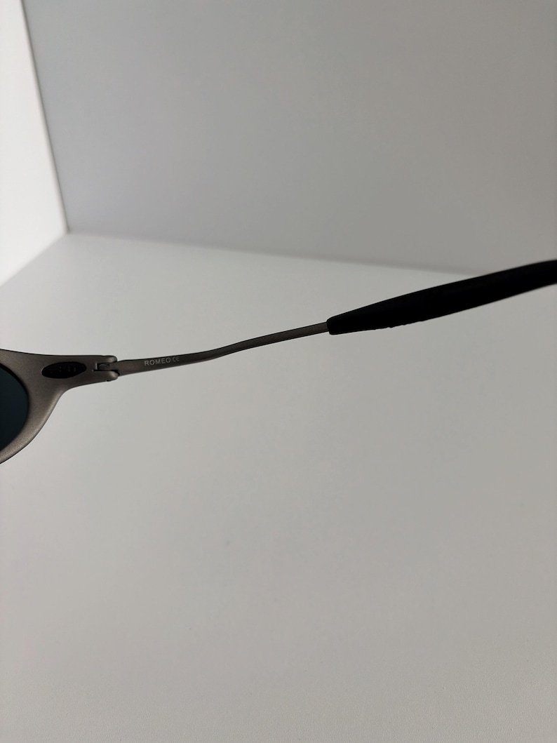 May include: Close-up of a pair of sunglasses with a matte gray frame and dark lenses. The temples are thin and curve towards the ear, with black rubberized tips for comfort. The text "ROMEO" is visible on the frame.