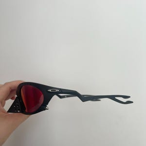 Może przedstawiać: Czarne okulary przeciwsłoneczne z czerwonymi soczewkami. Okulary mają unikalny design z tr&oacute;jkątnym akcentem z boku i elegancką, nowoczesną oprawką. Soczewki są w żywym czerwonym kolorze.