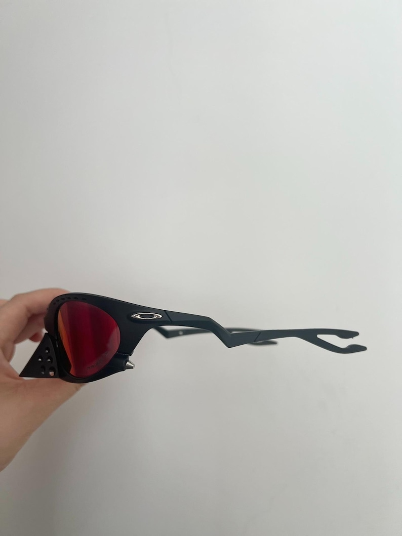 Może przedstawiać: Czarne okulary przeciwsłoneczne z czerwonymi soczewkami. Okulary mają unikalny design z tr&oacute;jkątnym akcentem z boku i elegancką, nowoczesną oprawką. Soczewki są w żywym czerwonym kolorze.