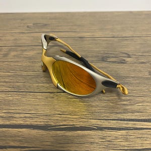 Puede incluir: Gafas de sol doradas y plateadas con lentes naranjas. Las gafas tienen un dise&ntilde;o elegante y moderno con una montura curvada y un puente nasal negro. Las lentes son de color naranja vibrante.