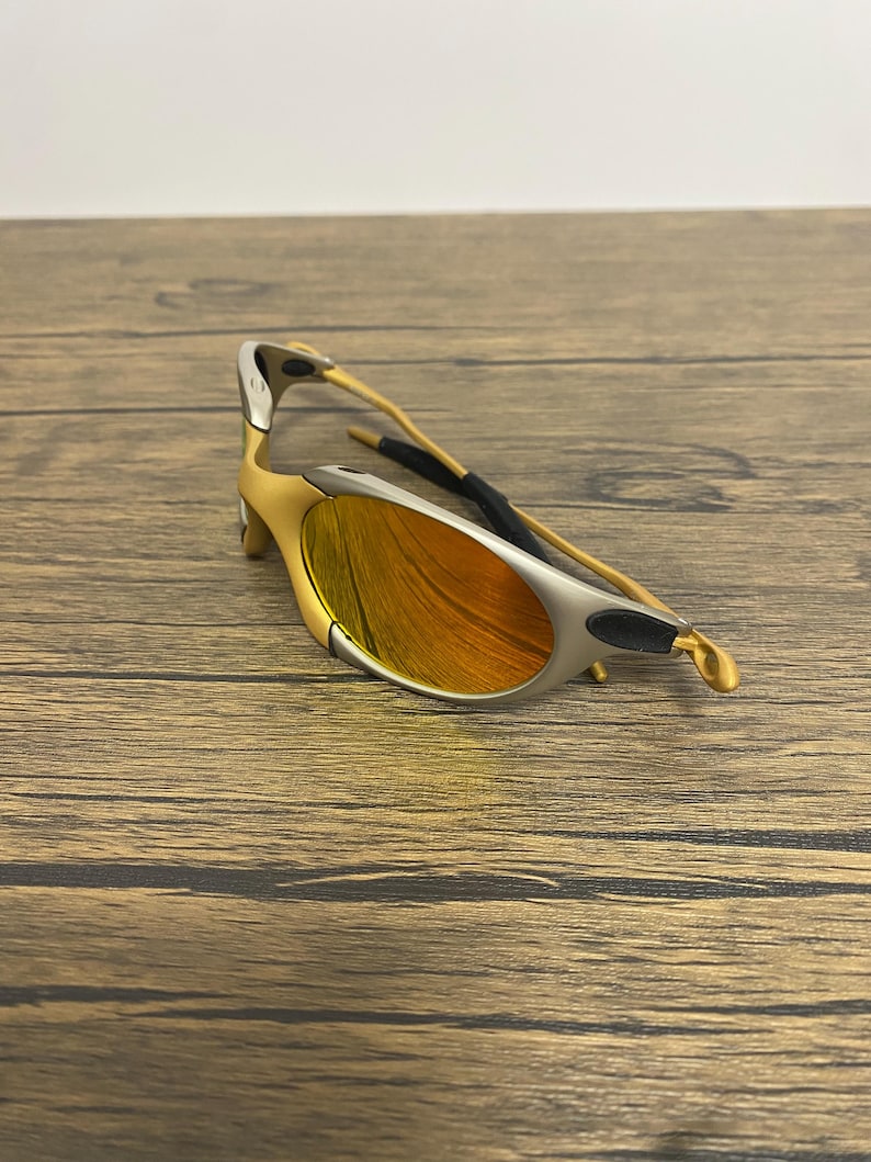 Puede incluir: Gafas de sol doradas y plateadas con lentes naranjas. Las gafas tienen un dise&ntilde;o elegante y moderno con una montura curvada y un puente nasal negro. Las lentes son de color naranja vibrante.