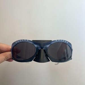 May include: Dark blue sunglasses with dark lenses. The sunglasses have a distinctive design, including a nose bridge and a curved frame. The frame features small, evenly spaced holes along the top edge. The sunglasses are held by a hand.