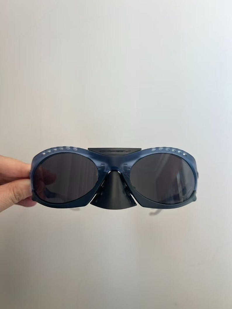 May include: Dark blue sunglasses with dark lenses. The sunglasses have a distinctive design, including a nose bridge and a curved frame. The frame features small, evenly spaced holes along the top edge. The sunglasses are held by a hand.