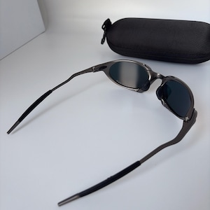 May include: Gunmetal sunglasses with dark lenses and a black protective case. The sunglasses have a sleek, modern design with curved temples and a matte finish. The case is oval-shaped and has a zip closure.