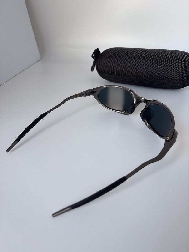 May include: Gunmetal sunglasses with dark lenses and a black protective case. The sunglasses have a sleek, modern design with curved temples and a matte finish. The case is oval-shaped and has a zip closure.
