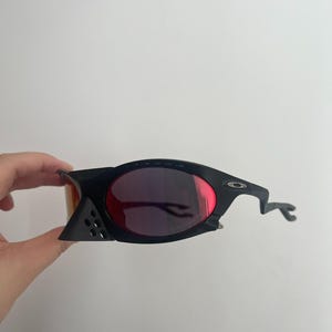 Może przedstawiać: Czarne okulary przeciwsłoneczne z czerwonymi soczewkami. Okulary mają unikalny, kanciasty design z zakrzywionym zausznikiem. Soczewki są w kolorze głębokiej czerwieni, a oprawki czarne.